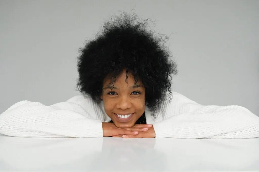 Smiling woman in a white sweater resting her chin on her hands against a light gray background.