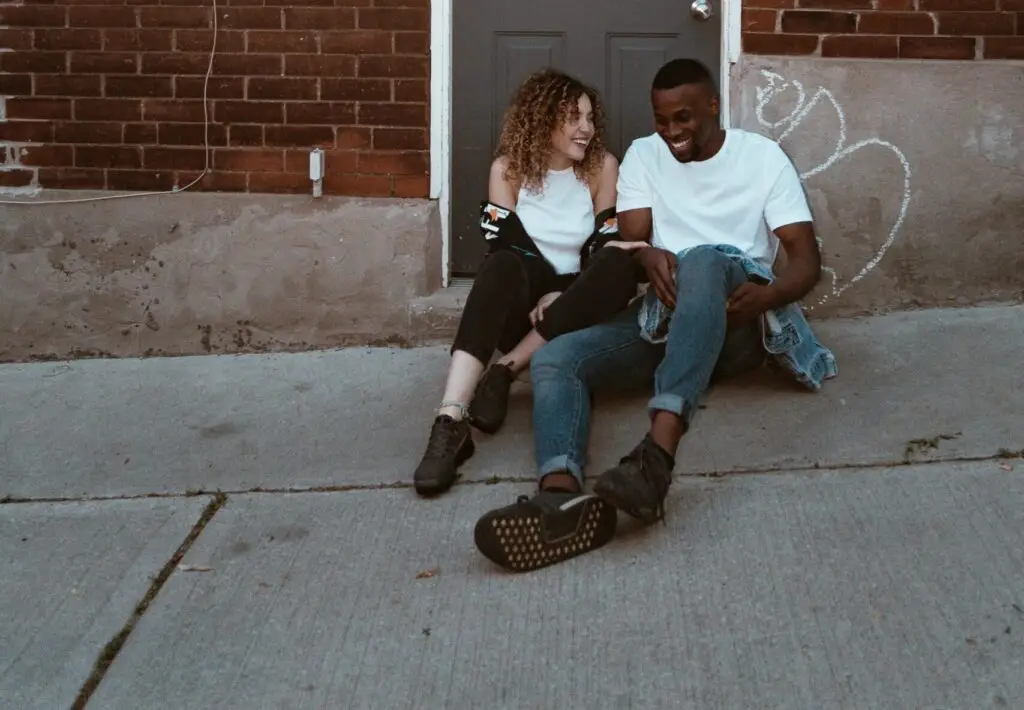 Two young adults sitting on a sidewalk against a brick wall, smiling and laughing together.