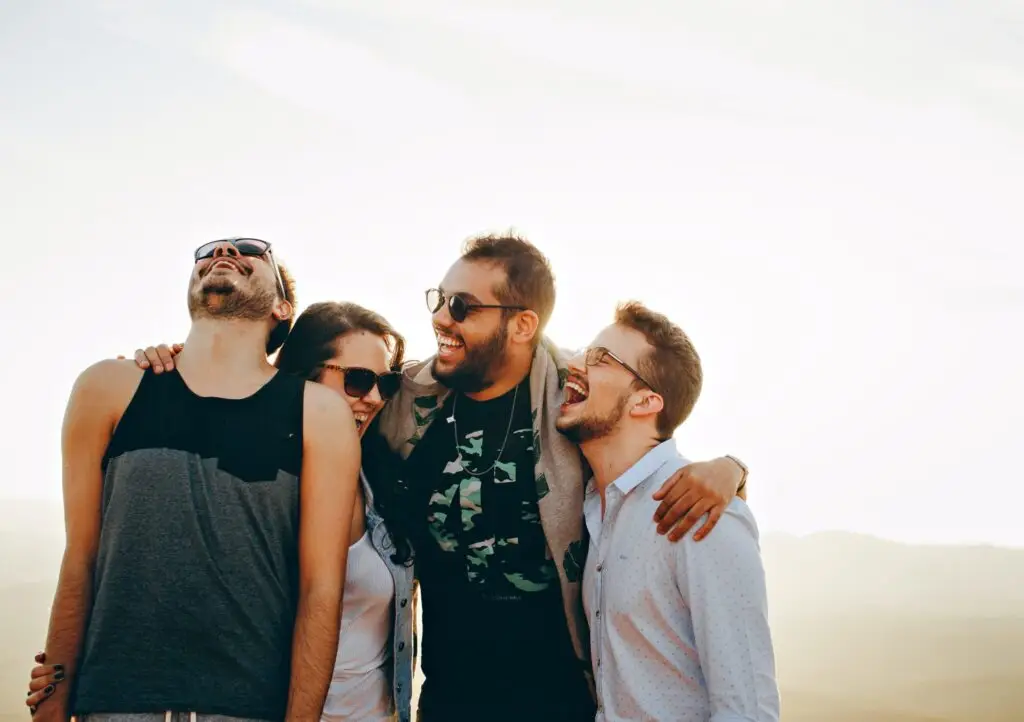 Four friends laughing together with arms around each other outdoors in bright sunlight.