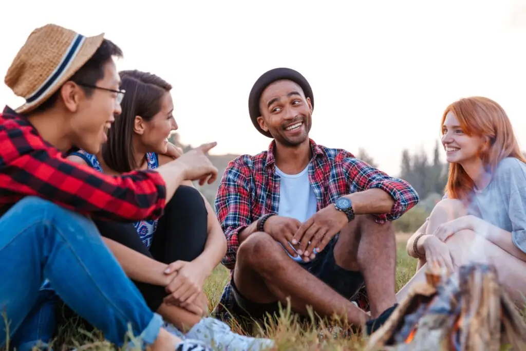 A diverse group of smiling young adults sitting together outdoors on grass, talking around a campfire at sunset.