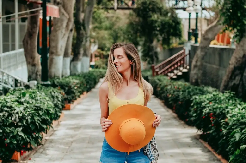 Smiling woman holding an orange sun hat while walking along a sunlit garden pathway lined with greenery.