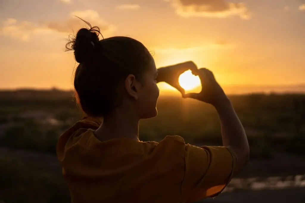 Woman forming a heart shape with her hands around the setting sun at golden hour, symbolizing hope and recovery.