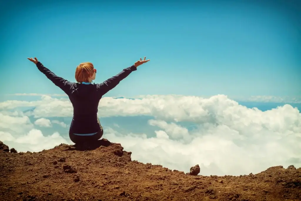Woman sitting on a mountain summit with arms raised above the clouds, embracing freedom and hope.