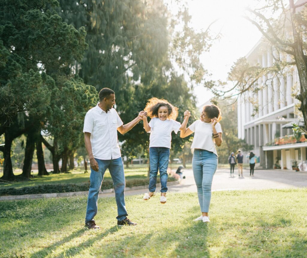 Smiling parents swing their laughing daughter by the hands in a sunny park, enjoying a joyful family moment together.