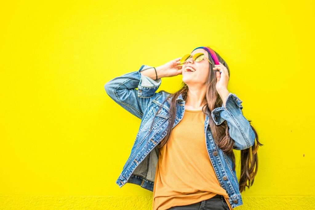 Smiling young woman in denim jacket and sunglasses laughing against a bright yellow wall.