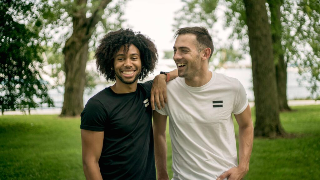 Two smiling men walking together outdoors in a green park, arm around shoulder, enjoying supportive friendship.