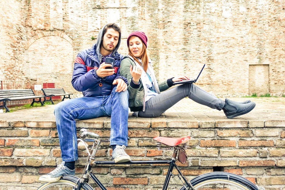 Teenage couple using phone and notebook outdoor at winter time - Best friends looking internet content sitting on old urban wall - Concept of young people lifestyle with modern technologies and trend